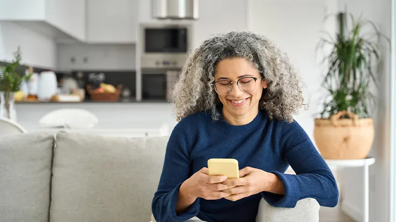 A woman with gray hair sits on a coach smiling and looking at her phone.